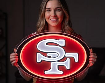 Può includere: Un'insegna a LED di forma ovale con il logo dei San Francisco 49ers in bianco, su uno sfondo rosso vibrante illuminato. L'insegna ha un bordo in acrilico trasparente ed emette una luce brillante, mettendo in risalto le iconiche lettere SF.