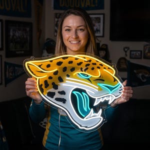 May include: A neon sign shaped like a jaguar head in profile, featuring yellow, black, white, and teal light tubes. The design depicts a snarling jaguar with black spots, a white muzzle, and teal accents on the eye and mouth, mounted on a clear backing.