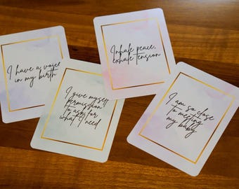 May include: Four white affirmation cards with gold borders and handwritten text. The cards have watercolor-style pink and purple accents. The phrases include "I have a voice in my birth," and "I am so close to meeting my baby."
