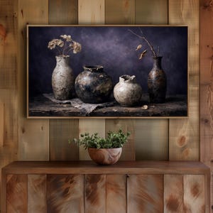 Pode incluir: Uma impressão emoldurada de natureza morta com quatro vasos texturizados de várias formas e tamanhos, dispostos sobre uma superfície de madeira rústica. Os vasos contêm flores secas e galhos, contra um fundo escuro e atmosférico. A impressão está acima de um armário de madeira.