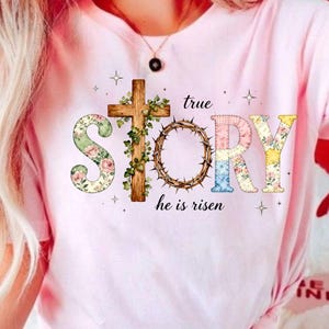 May include: A pale pink t-shirt with the words "true STORY he is risen" in a decorative font. The design includes a floral "S", a wooden cross, and a crown of thorns. The letters "T", "O", "R", and "Y" have floral patterns.
