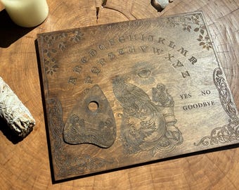 May include: A square wooden spirit board with an engraved alphabet, the words "As Above, So Below," "YES," "NO," and "GOODBYE," featuring a central raven and skeleton illustration, accompanied by a matching triangular planchette with a hole.