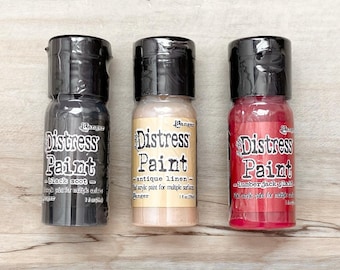 Puede incluir: Tres frascos pequeños de pintura Distress en negro, beige y rojo. Los frascos están etiquetados con "Distress Paint" y tienen un aspecto desgastado.
