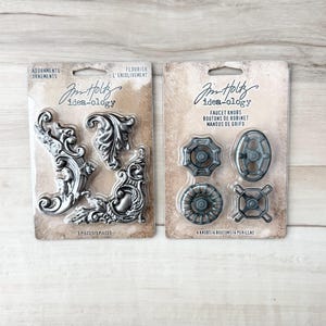 Puede incluir: Dos paquetes de adornos Idea-ology. Un paquete contiene piezas de esquina plateadas y ornamentadas. El otro paquete contiene cuatro pomos de grifo en varios diseños. Los paquetes están sobre una superficie clara de vetas de madera.