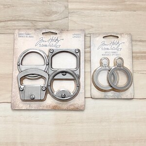 May include: Two packages of metal craft embellishments from the Tim Holtz Idea-ology line. The first package contains four metal frames, including a bottle opener and a handle. The second package contains two metal rings.