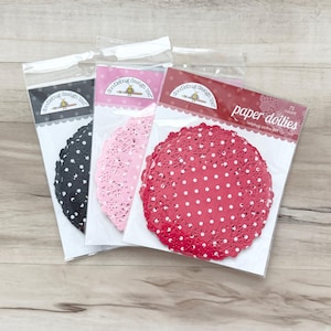 Op de afbeelding: Drie verpakkingen papieren onderzetters van Doodlebug Design Inc. De verpakkingen bevatten ronde, kanten onderzetters in zwart, lichtroze en rood, elk met een wit stippenpatroon. De rode verpakking is gelabeld met 75 count ladybug polka dot.