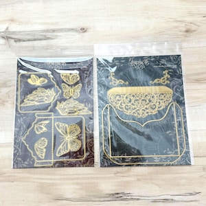 May include: Two clear plastic sleeves containing gold-colored metal cutting dies. The left sleeve features butterfly shapes and a small envelope die. The right sleeve contains a large, ornate, decorative envelope die with intricate filigree patterns.