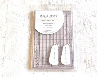 Puede incluir: Un cortador rápido Spellbinders Paper Arts Quick Trimmer en un paquete de plástico transparente. El cortador es gris y tiene una regla con pulgadas y centímetros. El paquete incluye dos herramientas de puntuación de plástico blanco con el logotipo de Spellbinders Paper Arts.