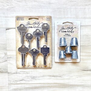 May include: Two packages of vintage-style metal embellishments for crafting. The first package contains seven keys with words like "Dream", "Found", "Lucky", "Fly", "Heart", "Brave", and "Curious". The second package contains seven silver thimbles with the words "Thimble" and "Des a Coudre" on the packaging.