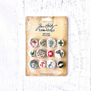 May include: A package of 12 mini flair buttons with various designs, including a clock, a butterfly, a star, and a flower. The package is labeled "Jim Holtz idea-ology Mini Flair Petit Flair 12 Pieces/12 Pieces".
