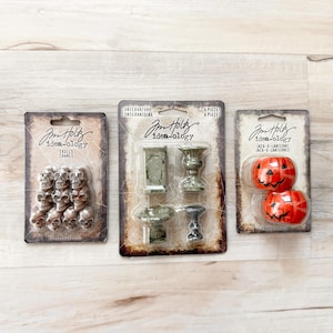 May include: Three packages of Tim Holtz Idea-ology embellishments. The packages contain skulls, miniature conservatory pieces, and Jack-o-Lanterns. The skulls are silver, the conservatory pieces are gray, and the Jack-o-Lanterns are orange.