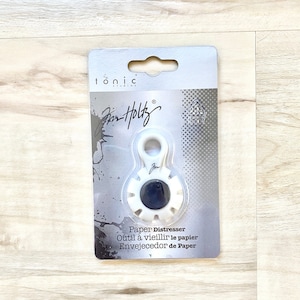 Puede incluir: Herramienta de envejecimiento de papel de plástico blanco con un centro negro y una perilla en la parte superior. El empaque dice "Paper Distresser, Outil à vieillir le papier, Envejecedor de Paper".