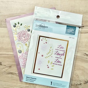 May include: A Spellbinders Rose Flourish cut and emboss folder in clear plastic packaging. The insert shows a white card with embossed roses, green leaves, and the text Live Every Moment, Laugh Every Day, Love Beyond Words in purple ink.