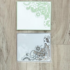 May include: A rectangular metal cutting die featuring an intricate corner design with gear-like and scrollwork patterns, packaged in a clear plastic sleeve, positioned above a sample cardstock cutout showing the same ornate corner motif.