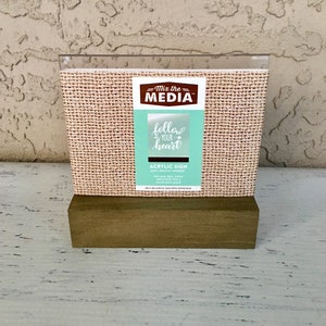 Op de afbeelding: Een bruine houten basis met een helder acryl bord waarop "Mix the MEDIA" en "follow your heart" in turquoise staat. Het bord is op een jute achtergrond.