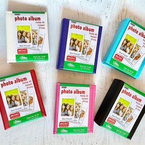 May include: Six photo albums in different colors, each with a photo of three friends on the front. The albums are all labeled "photo album" and say "Holds 40 Instant Prints". The albums are in white, blue, light blue, red, pink, and black.