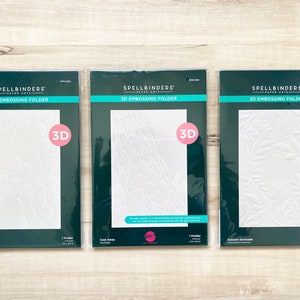 Puede incluir: Tres carpetas de relieve 3D Spellbinders. Las carpetas están empaquetadas en plástico transparente y tienen un fondo verde con texto blanco. Las carpetas están etiquetadas como "Floating Balloons", "Cast Away" y "Autumn Serenade".