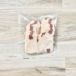 May include: A clear plastic bag filled with a stack of blank, cream-colored gift tags. The tags have a brown string attached to a hole at the top.