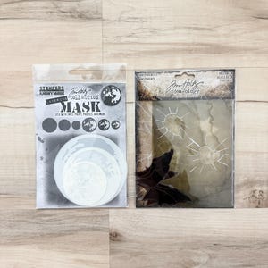 May include: Two craft supply packages on a light wood surface. One features a mask stencil with the text "MASK" and "STAMPERS ANONYMOUS". The other contains transparent sheets with a bat silhouette and the text "Jom Holtz idea-ology".