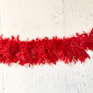 May include: A vibrant red, textured trim with a fluffy, fringed appearance. The trim is displayed against a white, weathered wooden background, creating a striking contrast. This decorative trim is suitable for various craft projects.