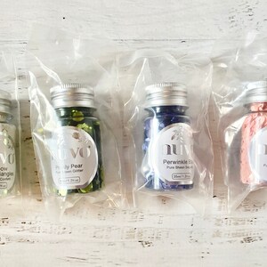 Puede incluir: Cuatro pequeños frascos de vidrio llenos de escamas de purpurina de colores. Cada frasco tiene una tapa plateada y una etiqueta con el nombre de la marca "NUVO" y el color y el nombre de la purpurina. Los colores son triángulos de cuarzo arcoíris, pera nacarada, azul periwinkle y tutú rosa.