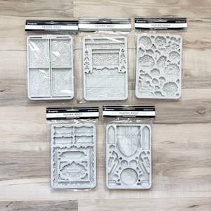 May include: A collection of five white silicone molds in clear packaging. The molds feature various decorative designs, including floral patterns, ornate frames, and architectural elements. Each mold is rectangular and designed for crafting.