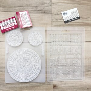 Puede incluir: Dos moldes de plástico transparente con diseños intrincados. Un molde presenta patrones circulares con números romanos, mientras que el otro tiene una variedad de formas rectangulares y cuadradas. El embalaje incluye etiquetas con el texto "TEXTURE IMPRESSION".