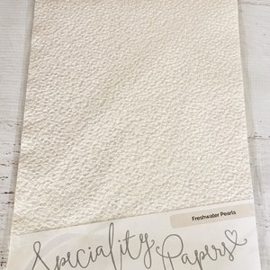 May include: A pack of 5 sheets of Freshwater Pearls A4 hand crafted cotton paper. The paper is white with a textured surface that resembles small pearls. The pack is labeled "Speciality Papers" and "Craft Perfect".