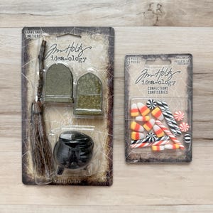 Puede incluir: Dos kits de manualidades con temática de Halloween. Uno incluye un caldero negro en miniatura, una escoba y dos lápidas. El otro contiene caramelos de maíz, bastones de menta y otros dulces. Ambos están empaquetados en plástico transparente.