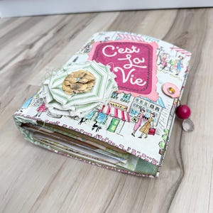 May include: A fabric-covered journal featuring a white background with a colorful illustration of a city street scene. A pink scalloped label with the text C'est la Vie is centered on the cover, accented by a layered green and white fabric flower.