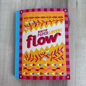 May include: A colourful book titled "flow" for paper lovers. The cover features a vibrant design with the text "BOOK FOR PAPER LOVERS" and "flow" in large, bold letters. The book has a blue spine and a red and pink checkered border.