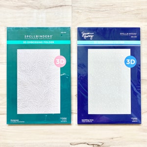 May include: Two 3D embossing folders for crafting. The folder on the left is green and features a white evergreen design. The folder on the right is blue and features a white snowflake design. Both folders are labeled "SPELLBINDERS' 3D EMBOSSING FOLDER".