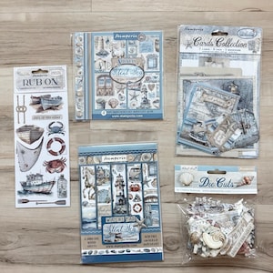 Puede incluir: Una colección de suministros para scrapbooking con temática náutica. Incluye hojas de pegatinas, troquelados, colecciones de tarjetas y washi pads. Los productos presentan imágenes de barcos, conchas marinas y faros. La paleta de colores es principalmente azul y blanco.