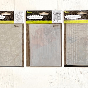 May include: Three clear plastic embossing folders for crafting. The folders are packaged in clear plastic with black and white text that reads "Darice Embossing Essentials 1PC/PZ 4.25in x 5.75in (10.7cm x 14.6cm)". The first folder has a pattern of diamonds, the second has a pattern of vertical lines, and the third has the word "thanks" repeated in a script font.
