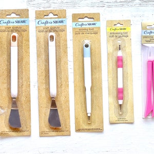 Könnte beinhalten: Fünf Bastelwerkzeuge in einzelnen Pappkartons verpackt. Die Werkzeuge umfassen einen Bastelspatel, ein Ritzwerkzeug, ein Prägewerkzeug und ein Set zum Stechen und Ritzen. Die Werkzeuge sind alle weiß mit rosa und blauen Akzenten.