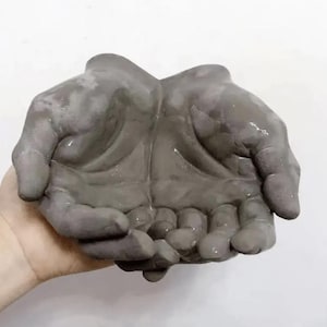 May include: A gray, hand-shaped decorative bowl. The bowl is designed to resemble two open hands, with the palms facing upward. The bowl is made of a smooth, matte material and is ideal for holding small items or as a decorative piece.