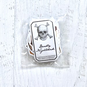 Puede incluir: Una pegatina blanca con un diseño de calavera y huesos cruzados. La pegatina dice "Deadly Nightshade" y "Extracted from the finest Atropa Belladonna foliage. Guaranteed to evoke the most bizarre delirium and hallucinations."