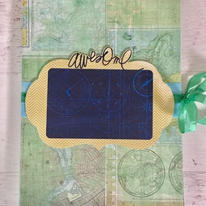 Puede incluir: Una cubierta de álbum de recortes con un patrón de chevron amarillo y azul, una brújula y la palabra "awesome" escrita en marcador negro. El fondo es un mapa con colores verde y azul.
