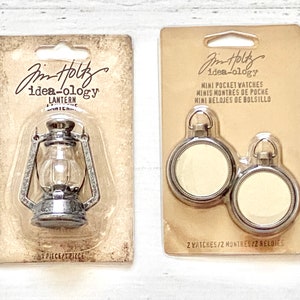 May include: Two packages of craft supplies from Jim Holtz idea-ology. The first package contains a silver metal lantern. The second package contains two silver pocket watches.