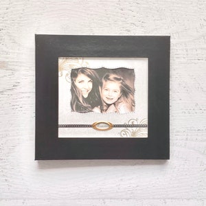 May include: Black picture frame with a white mat and a photo of two women, one with long brown hair and the other with long blonde hair. The frame has a gold ribbon with the word "Memories" on it.
