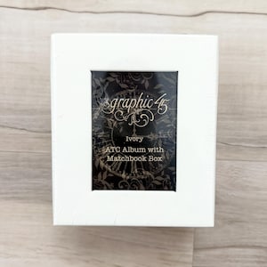 May include: Ivory-colored ATC album with a matchbook box. The album features a decorative black and gold design with the words "graphic 45" in an elegant script. The album is wrapped in clear plastic and sits on a light wood surface.