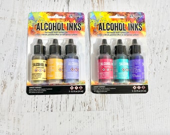 May include: Two sets of three alcohol inks in bottles with droppers. The inks are labeled "Alcohol Inks" and the colors are Lemonade, Flamingo, and Cosmic Blue. Each set is packaged in a clear plastic sleeve with a colorful background.