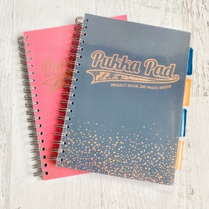 May include: Two spiral bound notebooks, one pink and one gray with gold accents. The gray notebook has the text "Pukka Pad PROJECT BOOK 200 PAGES 80GSM" printed on the front.