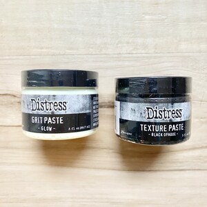 Puede incluir: Dos pequeños frascos de pintura artesanal de la marca Distress. El frasco de la izquierda es blanco y está etiquetado como "Grit Paste - Glow - 3 fl oz (86.7 ml)". El frasco de la derecha es negro y está etiquetado como "Texture Paste - Black Opaque - 3 fl oz (86.7 ml)".