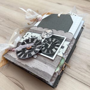 Puede incluir: Un álbum de recortes hecho a mano con un tema vintage. La portada presenta un diseño de vetas de madera gris, adornado con esferas de reloj, un adorno de mariposa y una etiqueta gris oscuro. El álbum está atado con una cinta y encaje blancos.