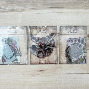 Puede incluir: Tres paquetes de efímeras Tim Holtz Idea-ology. Los paquetes contienen efímeras de papel con diseños vintage, incluyendo una mariposa, elementos florales y texto. El embalaje tiene una estética vintage y envejecida.