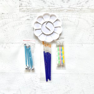 Puede incluir: Una paleta de pintura blanca en forma de flor con múltiples pozos, junto con varias herramientas de arte. Las herramientas incluyen herramientas de punteado con mango azul, pinceles con mangos azules y herramientas de punteado de doble punta con mangos coloridos. Los artículos están dispuestos sobre una superficie de madera blanca.