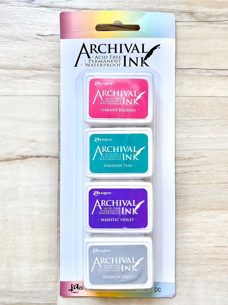 May include: A set of four ink pads in a clear plastic packaging. The ink pads are labeled "Ranger Archival Ink" and are in the colors vibrant fuchsia, paradise teal, majestic violet, and shadow grey.