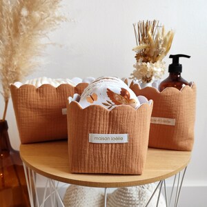 Peut inclure: Trois paniers de rangement en tissu couleur terre cuite avec des bords festonnés sur une table en bois. Les paniers contiennent des articles en tissu blanc et à motifs. Un panier porte l'étiquette "maison loëlle". Une bouteille en verre marron et des fleurs séchées sont également sur la table.