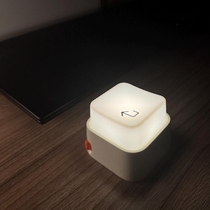 Può includere: Una piccola lampada quadrata e illuminata con una parte superiore bianca e una base grigio chiaro. La parte superiore ha un simbolo a freccia nera. La lampada è su una superficie di legno.